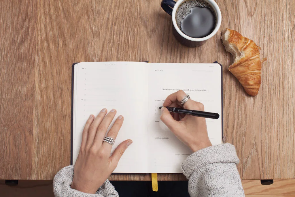 woman writing in journal on desk with coffee and croissant