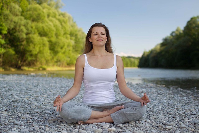 middle aged woman poses in yoga pose while sitting on rocks by a body of water in nature